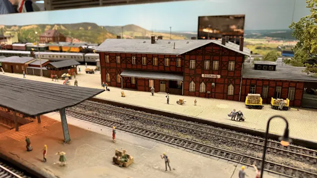 Tiny figures stand outside a large brick station building behind a covered platform.