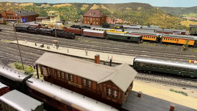 A small foreground station building faces a broad model railway yard and distant trains.