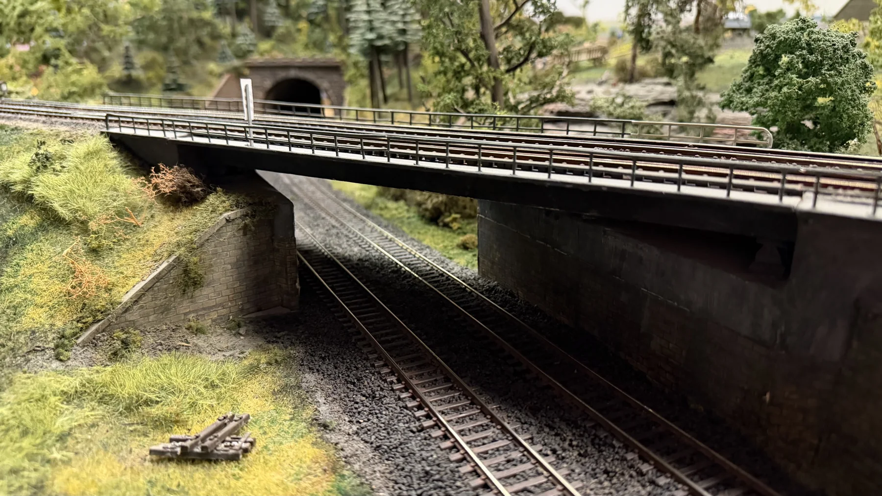 Model railway tracks lead into shadow beneath a road bridge with a metal railing.