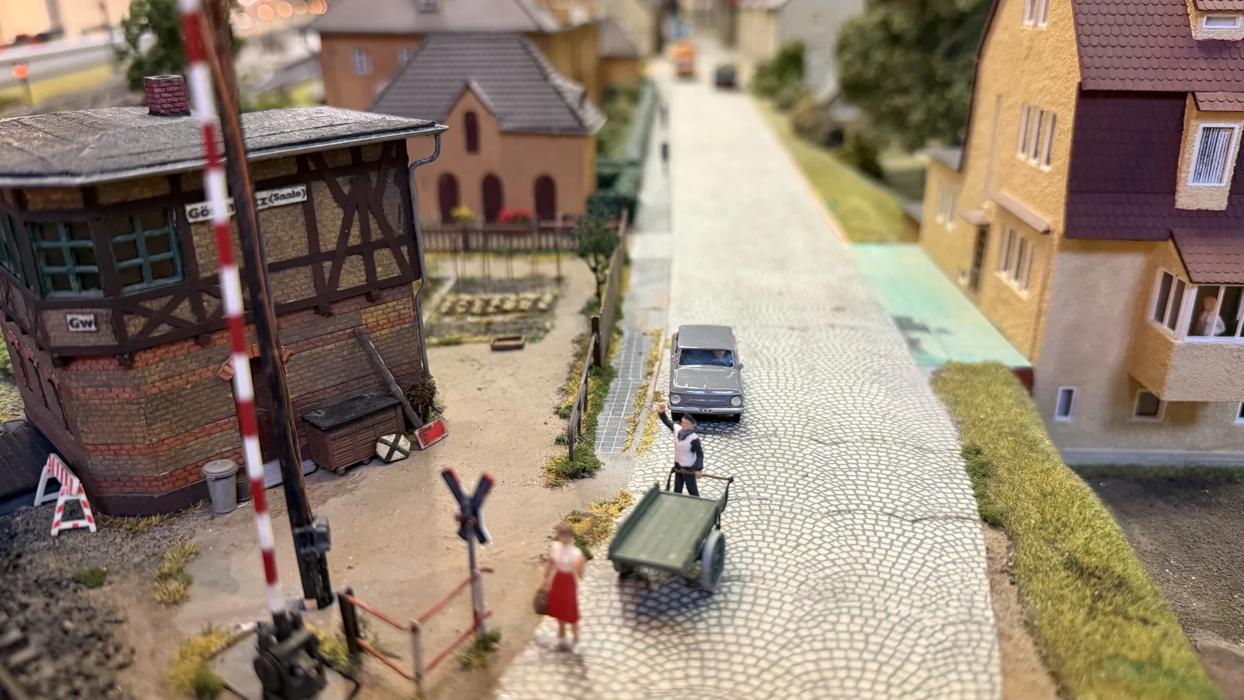 Small figures, a car and railway-side buildings line a cobbled model street.