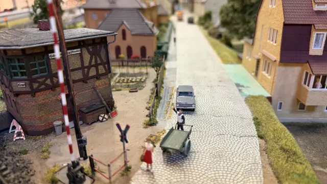Small figures, a car and railway-side buildings line a cobbled model street.