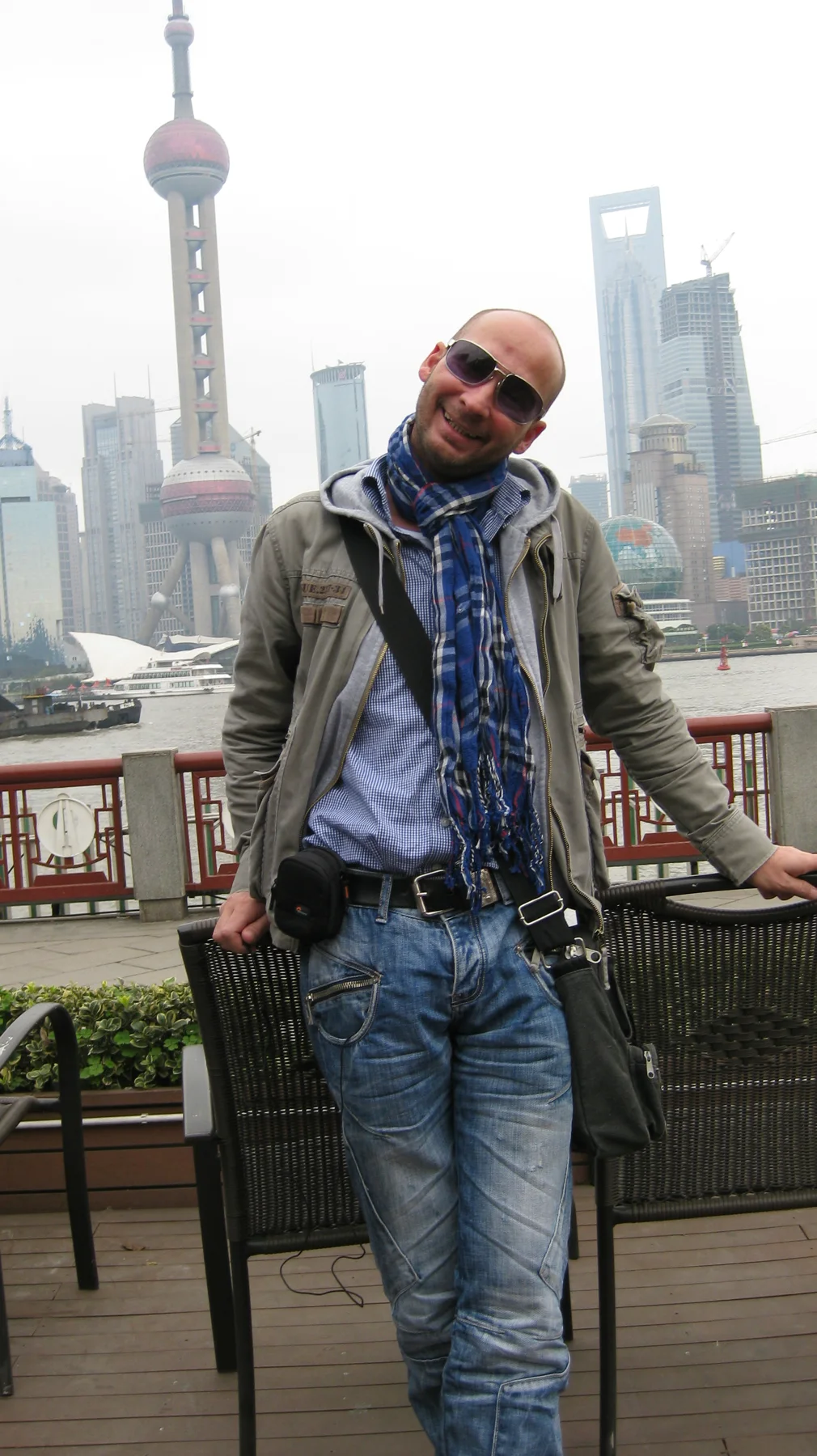 A person wearing sunglasses, a scarf and a jacket stands beside a railing, with tall buildings across the river behind.