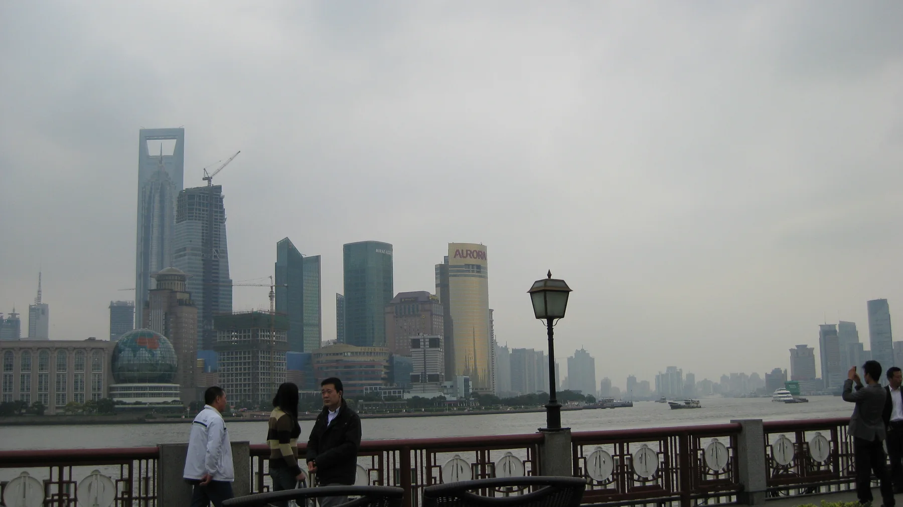 People stand beside a riverside railing and a dark lamp post, facing a hazy skyline of high-rise buildings.
