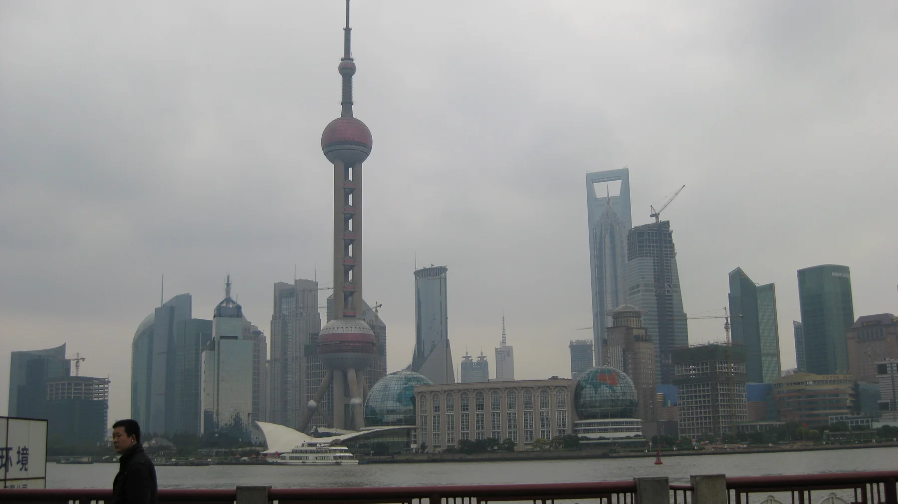 A slender tower with large spherical sections stands among skyscrapers across the river, with a railing in the foreground.