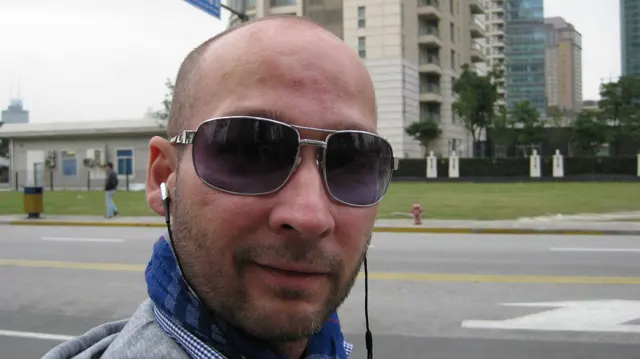 A person wearing sunglasses and earphones fills the foreground, with a road, lawn and tall buildings behind.