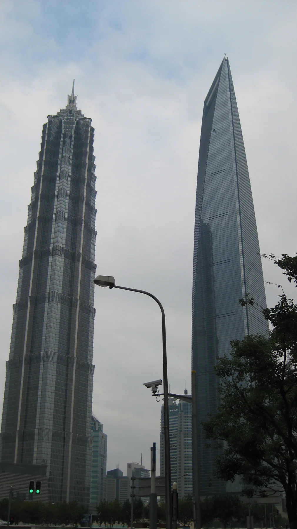 Two tall skyscrapers rise above a street lamp and treetops, one with a stepped facade and the other a smooth sloping profile.