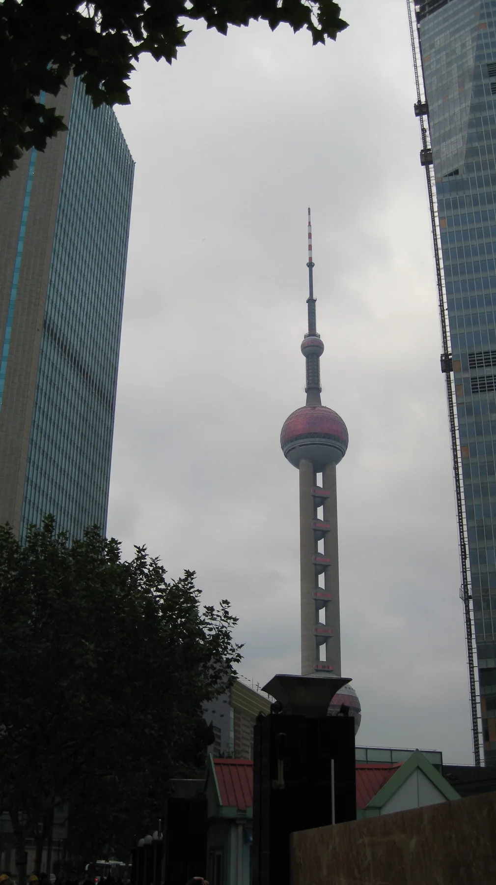 A slender tower with spherical sections is framed by high-rise facades and dark foliage in a portrait view from below.