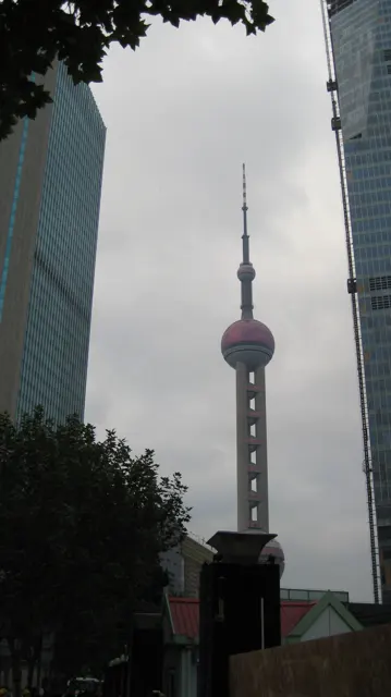 A slender tower with spherical sections is framed by high-rise facades and dark foliage in a portrait view from below.