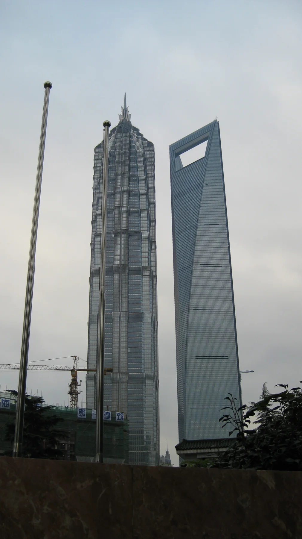 A stepped skyscraper and a tall building with a rectangular opening near its top rise side by side above trees and street furniture.