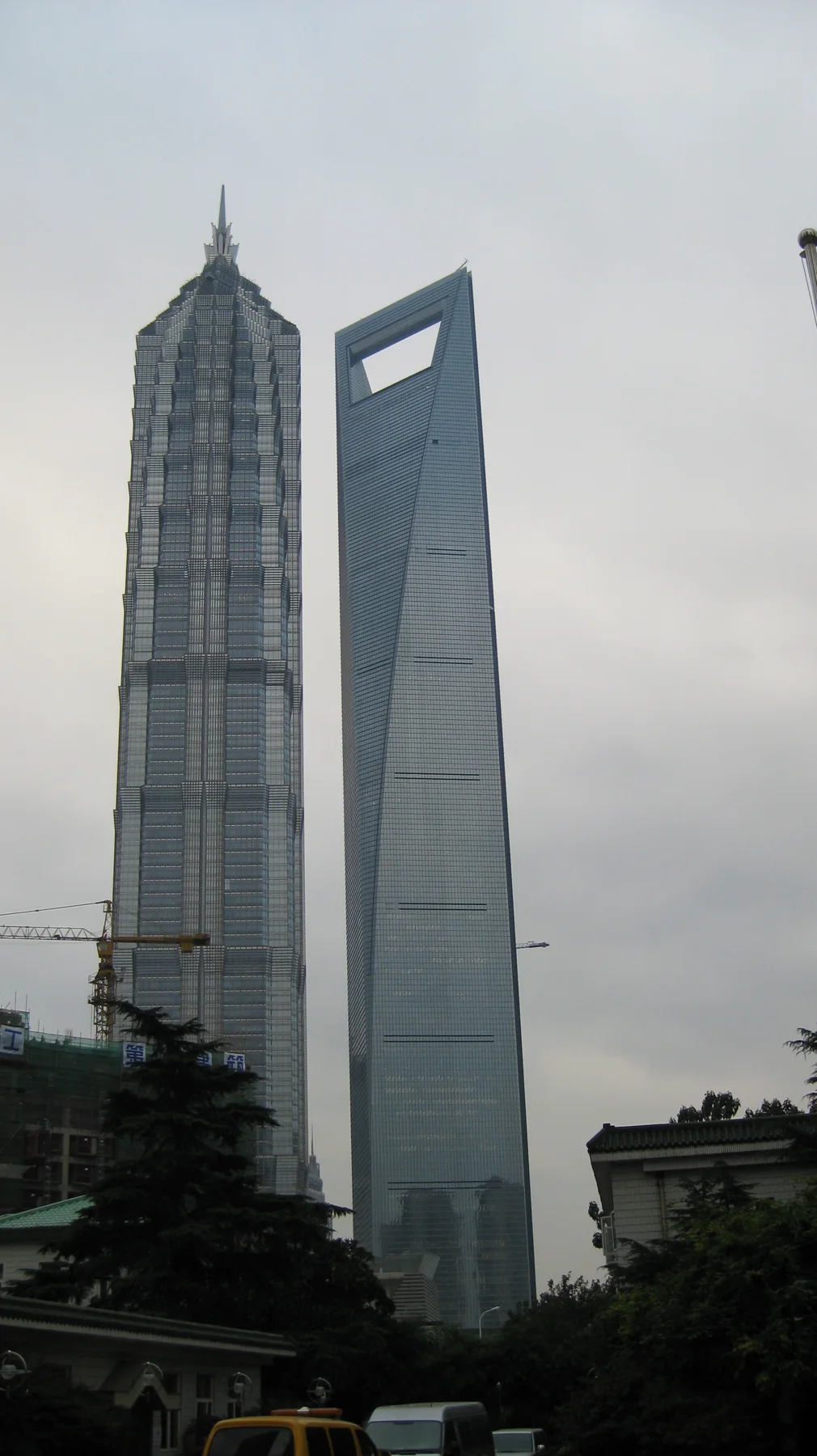 Two neighbouring skyscrapers dominate a portrait view beneath a pale overcast sky, with trees and vehicles below.