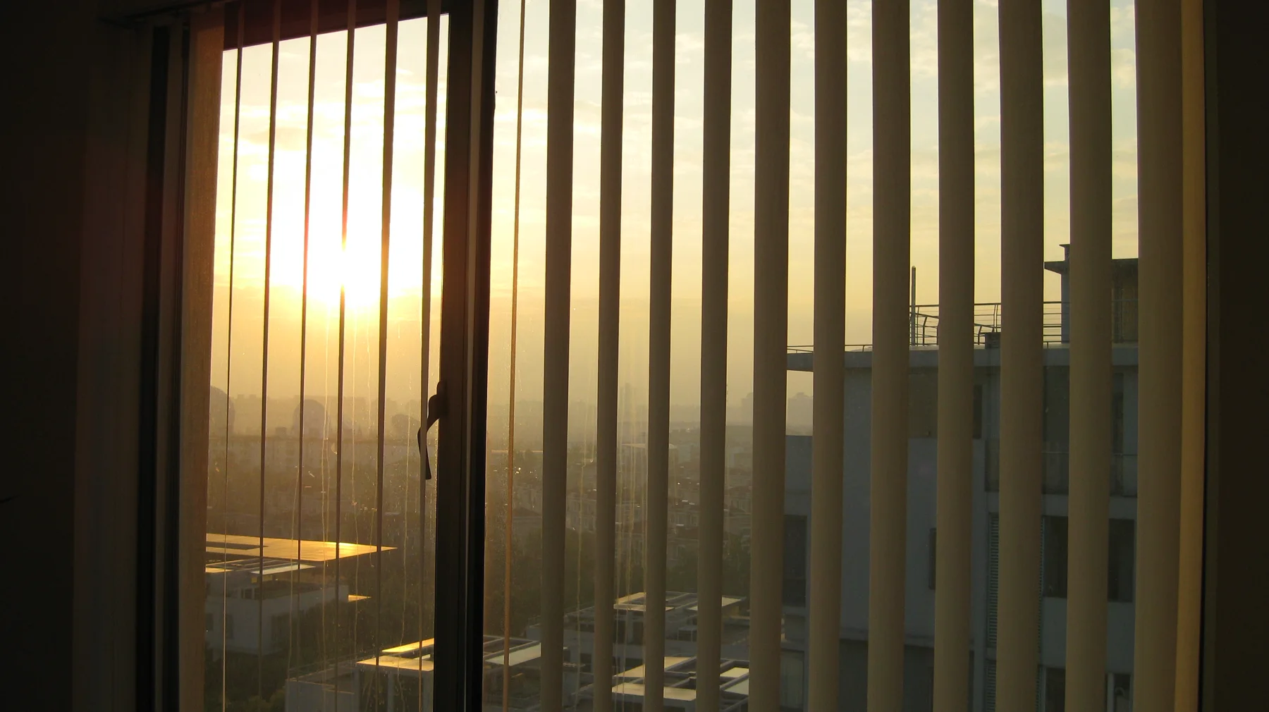 Low golden sunlight streams between vertical window blinds, with a hazy cityscape visible through the gaps.
