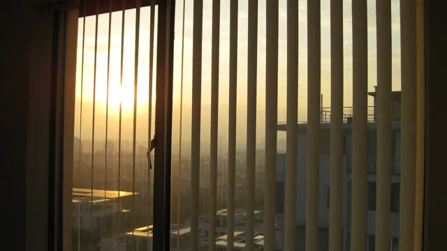 Low golden sunlight streams between vertical window blinds, with a hazy cityscape visible through the gaps.