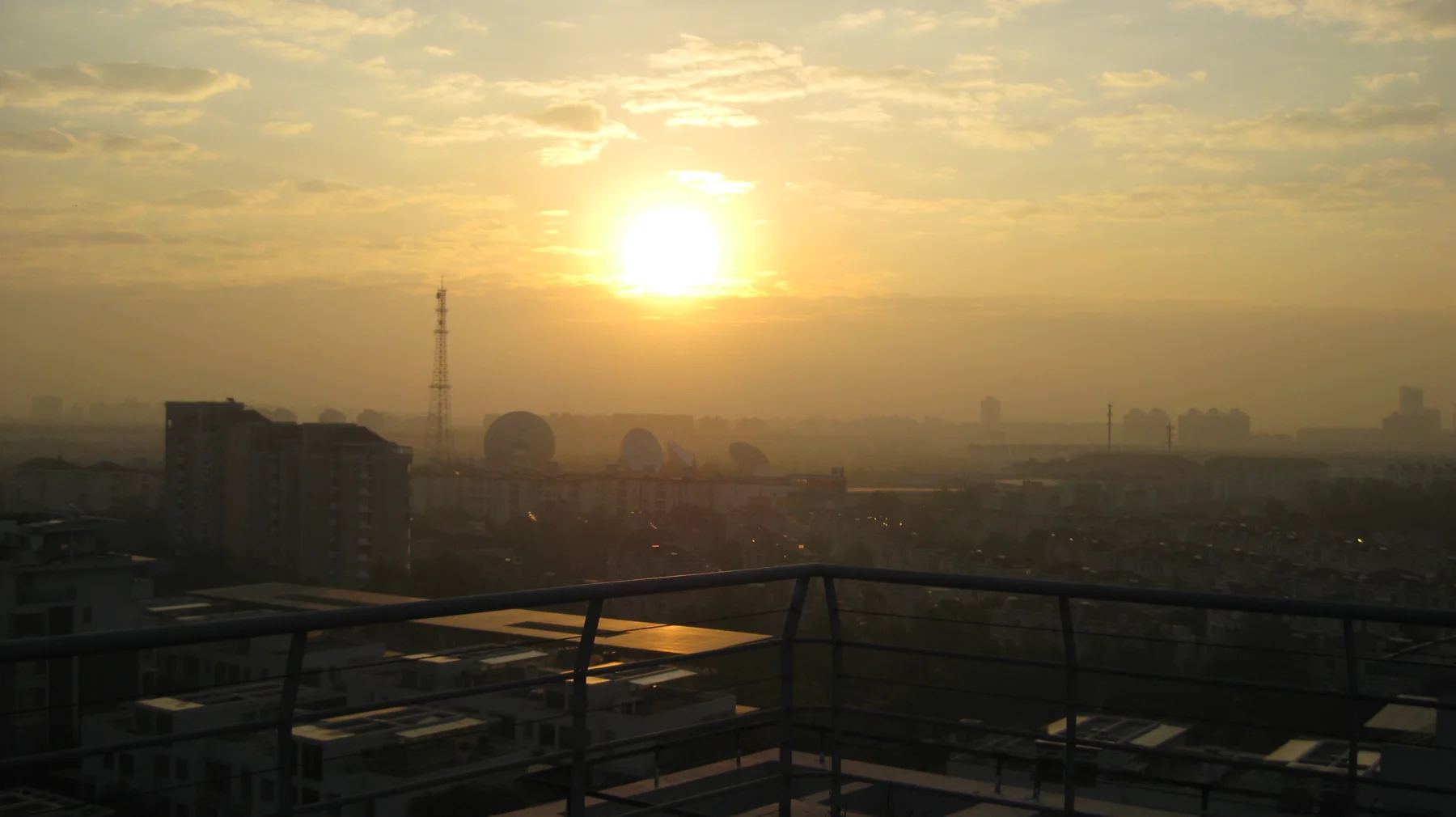A low bright sun hangs above a hazy spread of buildings beneath golden clouds, with a railing along the foreground.