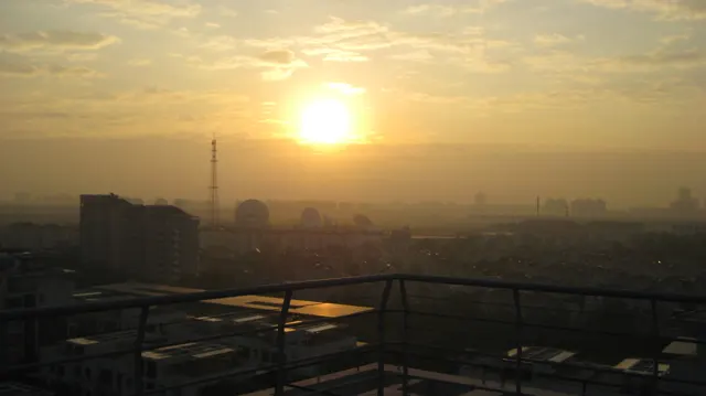 A low bright sun hangs above a hazy spread of buildings beneath golden clouds, with a railing along the foreground.