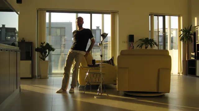 A person in a dark shirt and pale trousers stands beside armchairs in a warmly lit room, with open balcony doors behind.