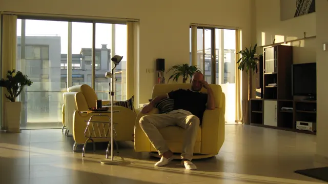 A person sits in a pale armchair in a sunlit room with large windows, plants and dark shelving.