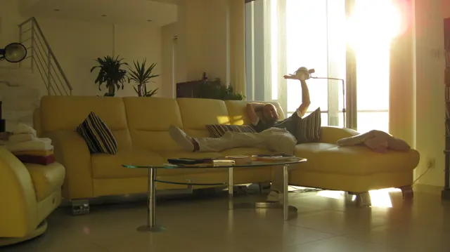 A person reclines on a pale sofa beside a glass-topped table, with bright sunlight entering through a tall window.