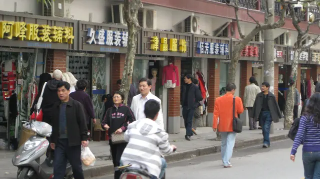 Pedestrians and scooter riders share a street lined with shops and Chinese signs, with bare tree trunks along the pavement.