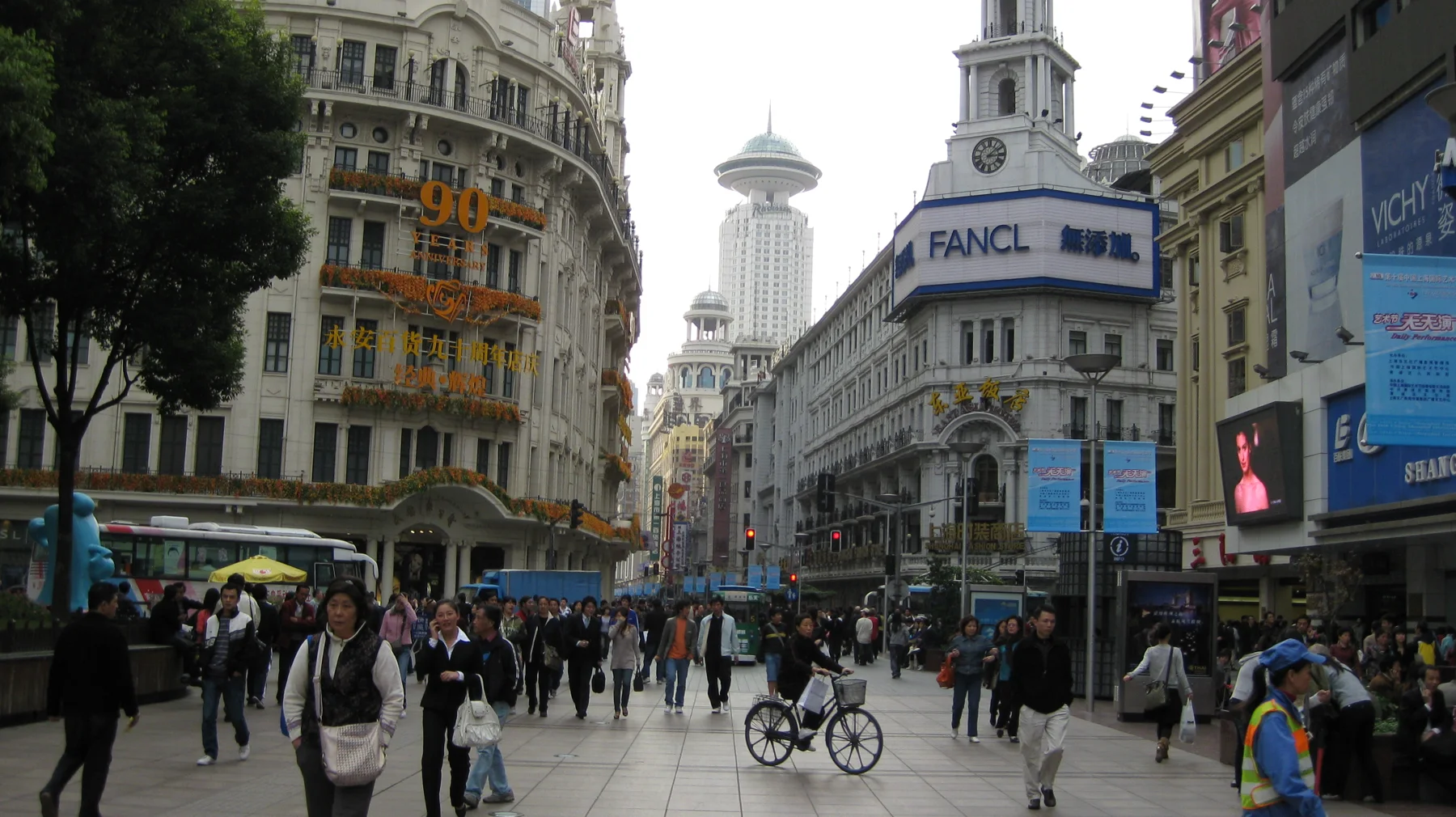 Pedestrians and cyclists move through a broad shopping street between tall buildings covered with signs and advertisements.