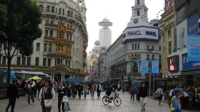 Pedestrians and cyclists move through a broad shopping street between tall buildings covered with signs and advertisements.
