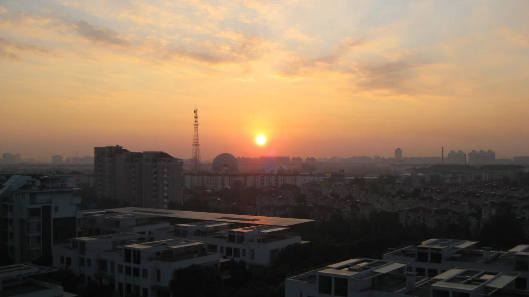 A low orange sun glows above rooftops and a distant tower beneath a warm, hazy sky.