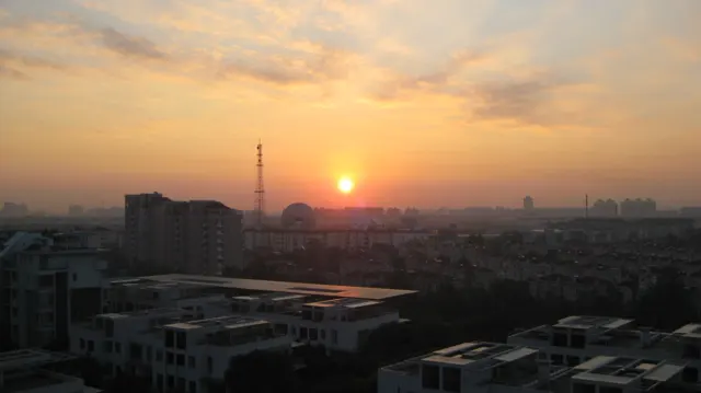 A low orange sun glows above rooftops and a distant tower beneath a warm, hazy sky.