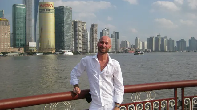 A person in a white shirt leans against a riverside railing, with water and a line of tall buildings in the background.
