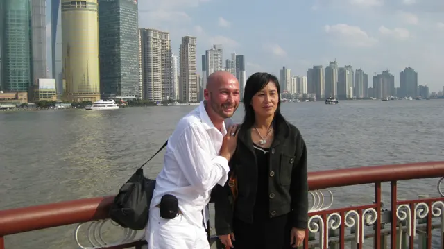 Two people pose beside a red-and-white railing, with the river and a high-rise skyline behind them.