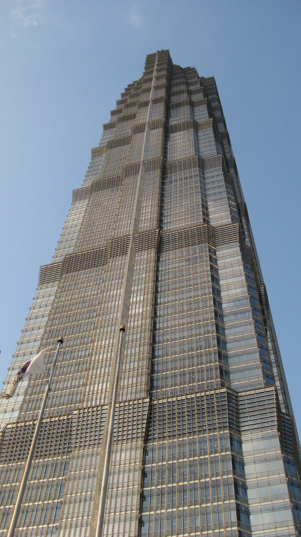 A close upward view follows the layered facade of a skyscraper towards its narrowing top against a blue sky.