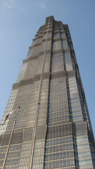 A close upward view follows the layered facade of a skyscraper towards its narrowing top against a blue sky.