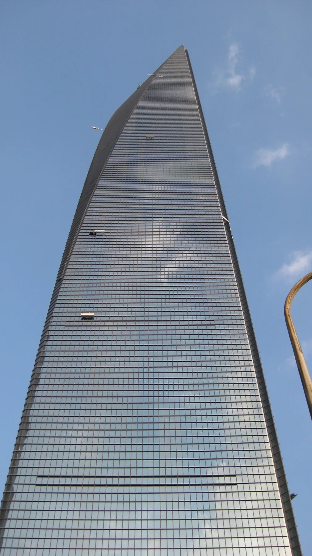 An upward view follows the smooth glass facade of a skyscraper towards a sharp sloping edge against the sky.