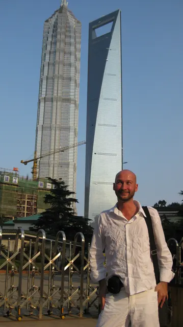 A person carrying a camera stands in the foreground beneath two tall skyscrapers, one stepped and one with an opening near its top.