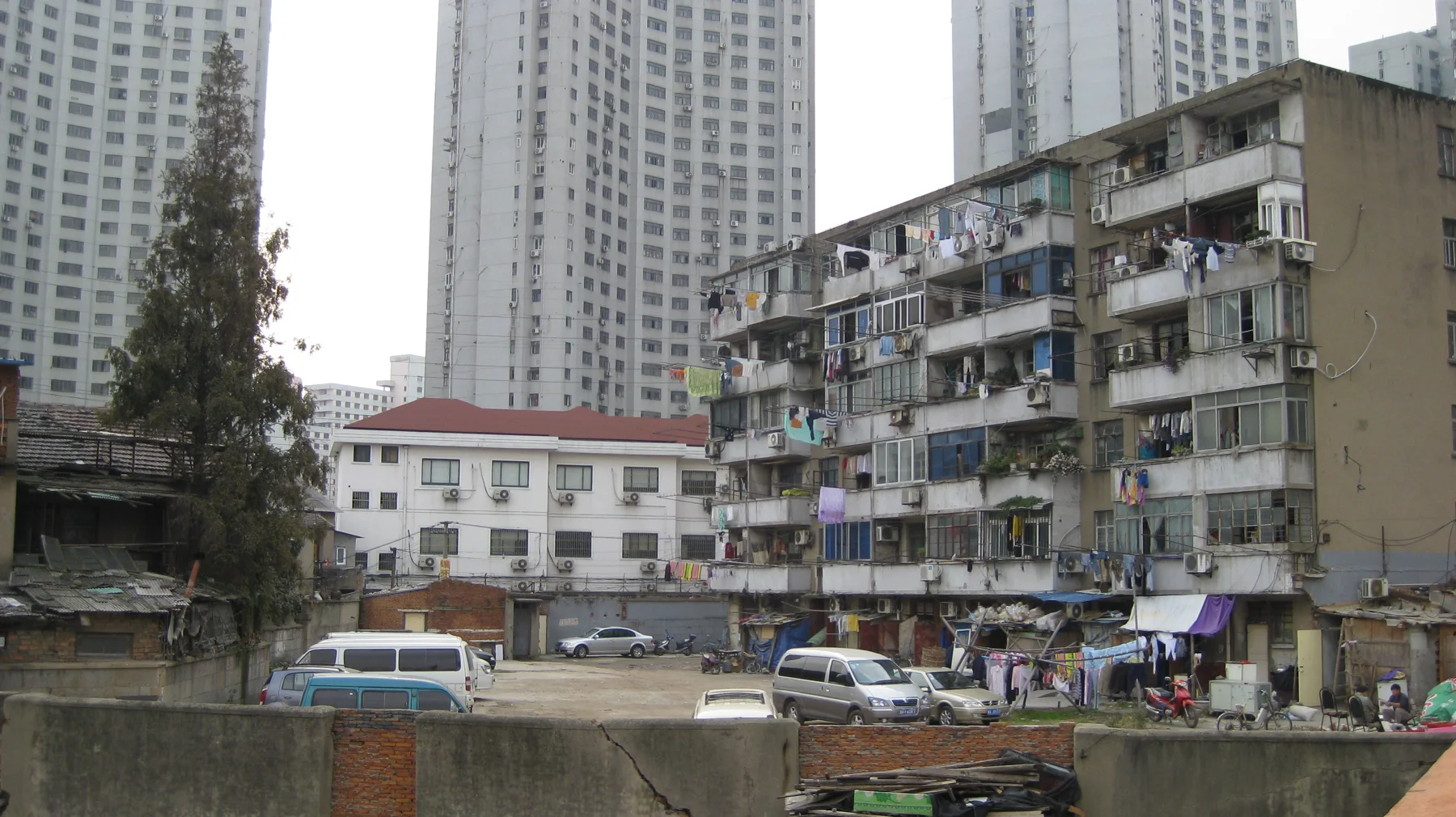 Low apartment blocks and parked vehicles stand beside a waterway, with much taller residential towers behind.