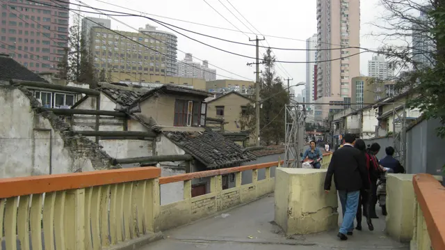 People walk over a small bridge with yellow-and-orange railings between low buildings and overhead wires.