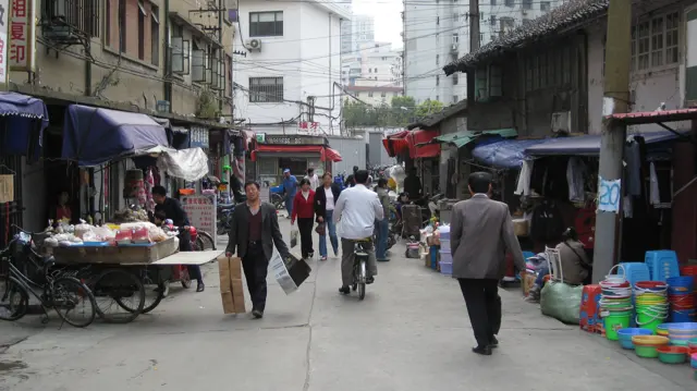 Pedestrians and a cyclist move along a narrow market street lined with stalls, awnings and stacked goods.