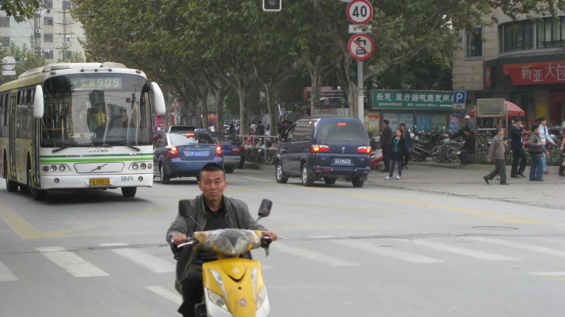 A rider on a yellow scooter approaches a road crossing, with a bus and other traffic behind.