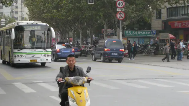 A rider on a yellow scooter approaches a road crossing, with a bus and other traffic behind.