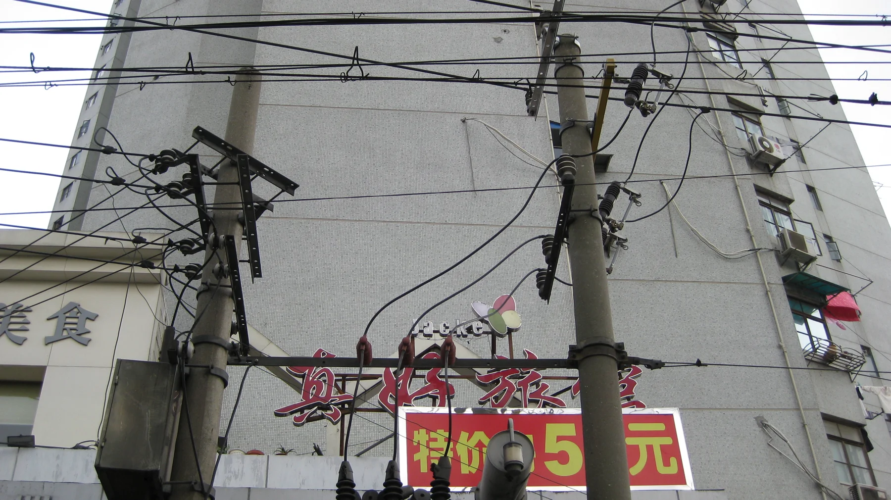 A dense web of utility wires runs between poles above shop signs and a pale building wall.