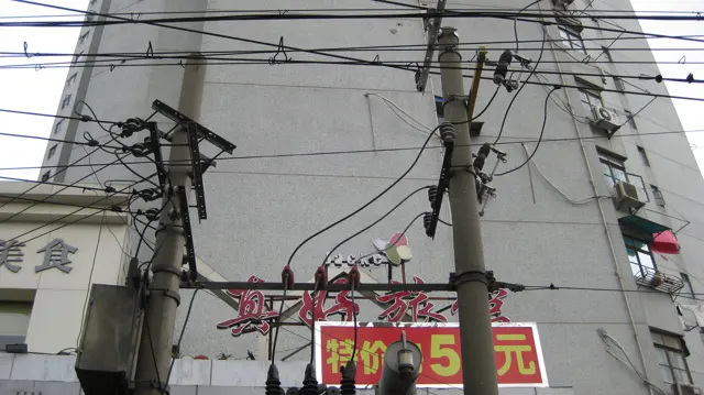 A dense web of utility wires runs between poles above shop signs and a pale building wall.