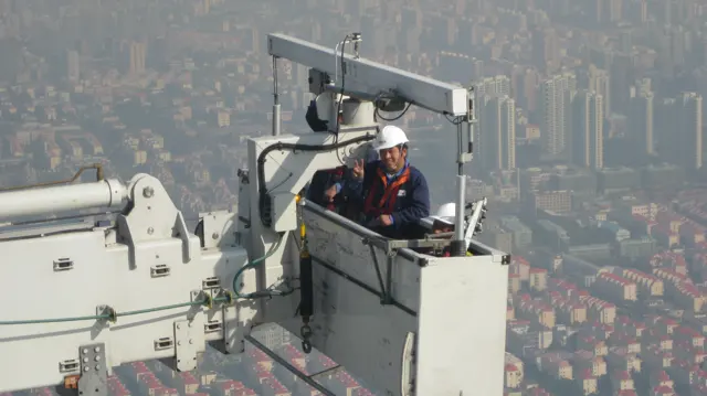 A helmeted worker stands in a suspended maintenance cradle with the hazy city far below.
