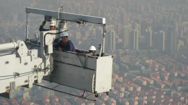 Two helmeted workers occupy a suspended platform beside metal machinery, high above the surrounding buildings.