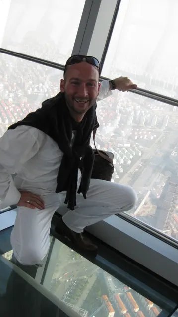 A person in pale clothing poses beside an observation window, with a hazy cityscape below.