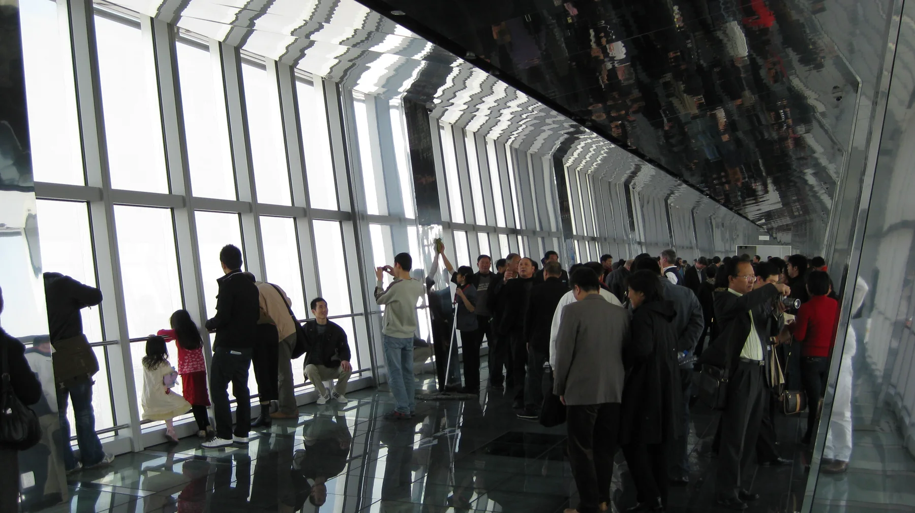 People gather beside tall angled windows beneath a dark reflective ceiling in a high observation corridor.