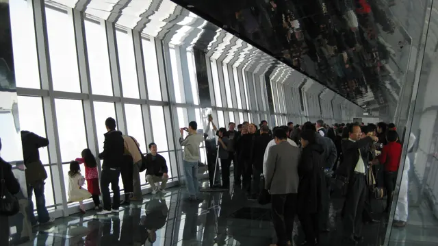 People gather beside tall angled windows beneath a dark reflective ceiling in a high observation corridor.