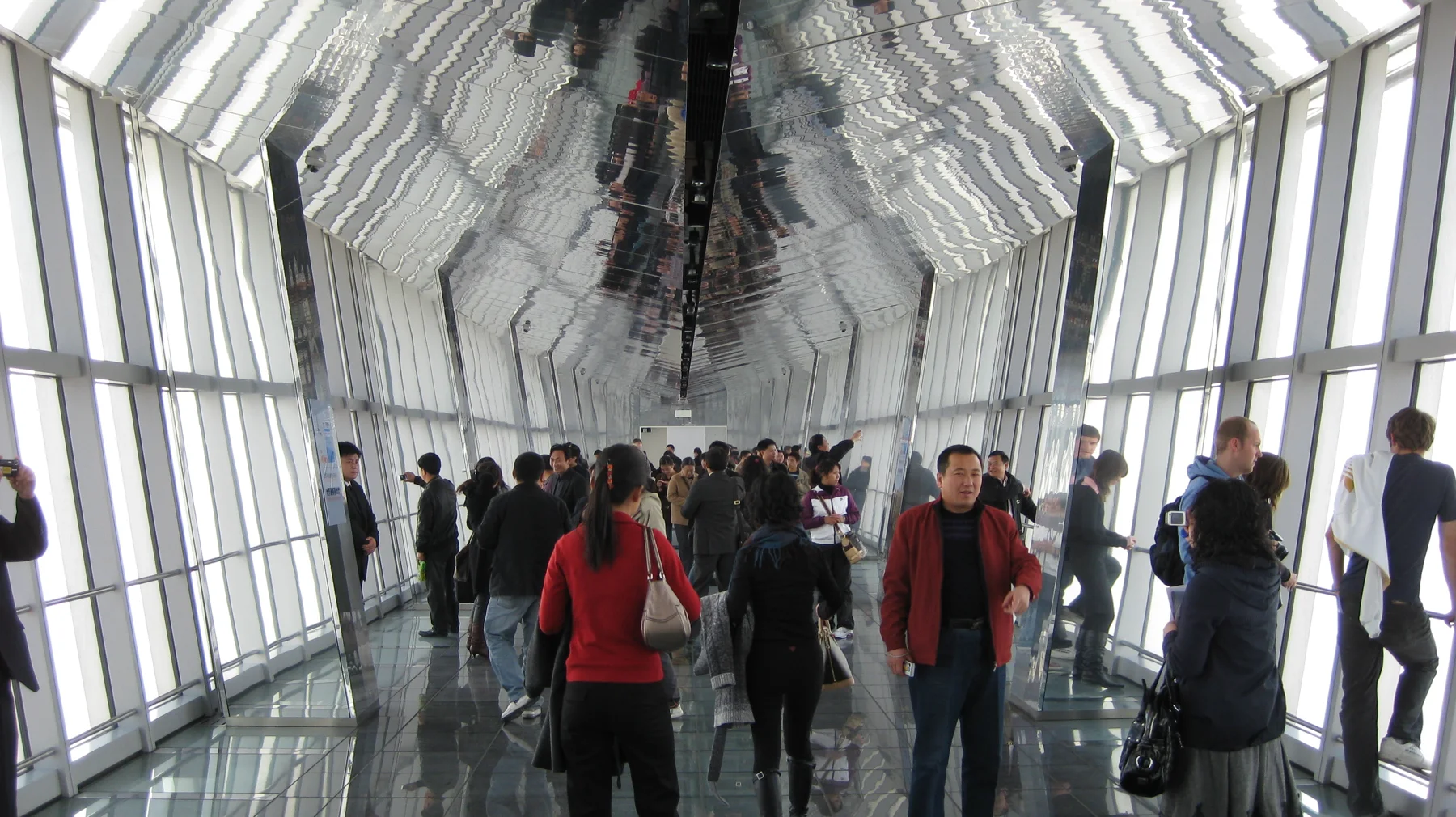 Visitors walk along a glass-sided observation corridor beneath a ceiling reflecting the windows and crowd.