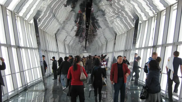Visitors walk along a glass-sided observation corridor beneath a ceiling reflecting the windows and crowd.