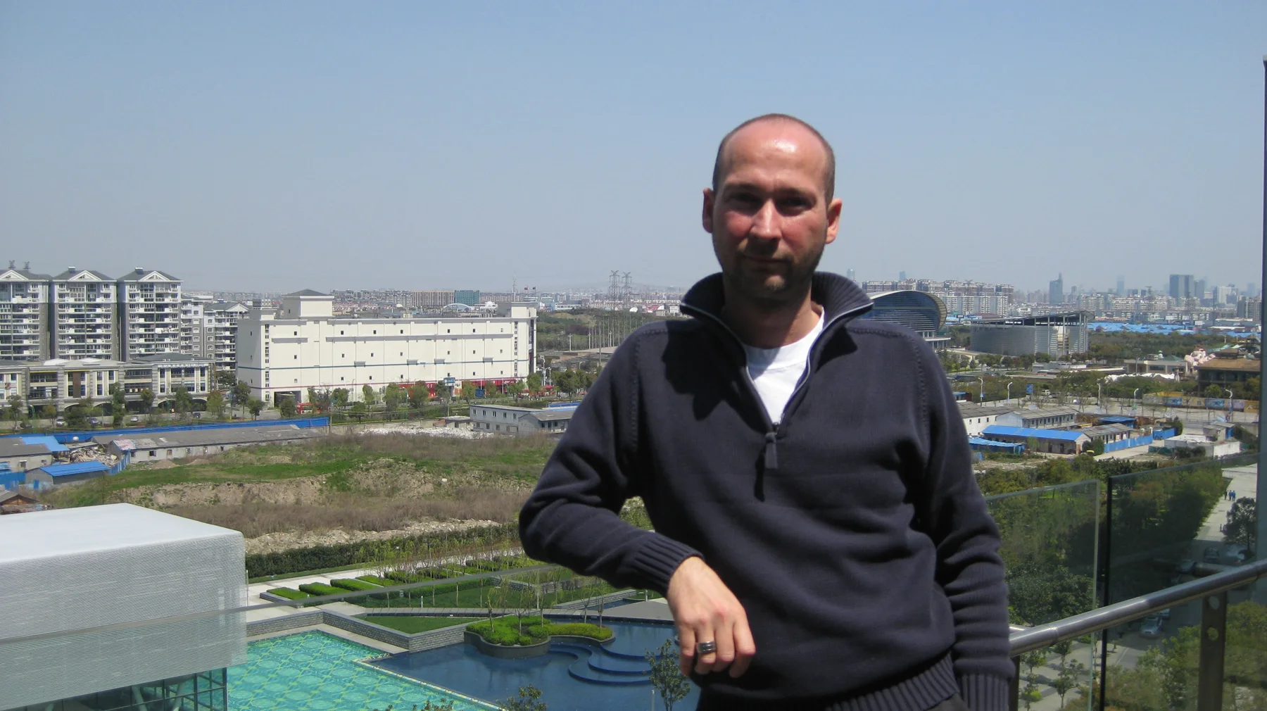 A person in a dark top leans against a glass balcony railing with buildings and open ground beyond.
