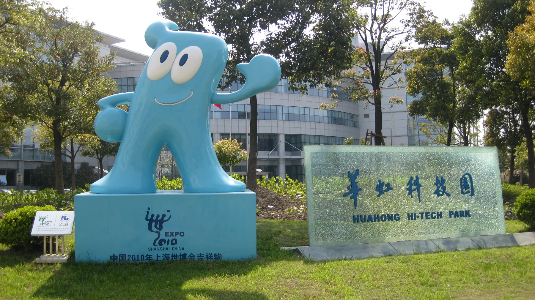 A large smiling blue mascot stands on a plinth beside a stone sign, surrounded by lawns and trees.