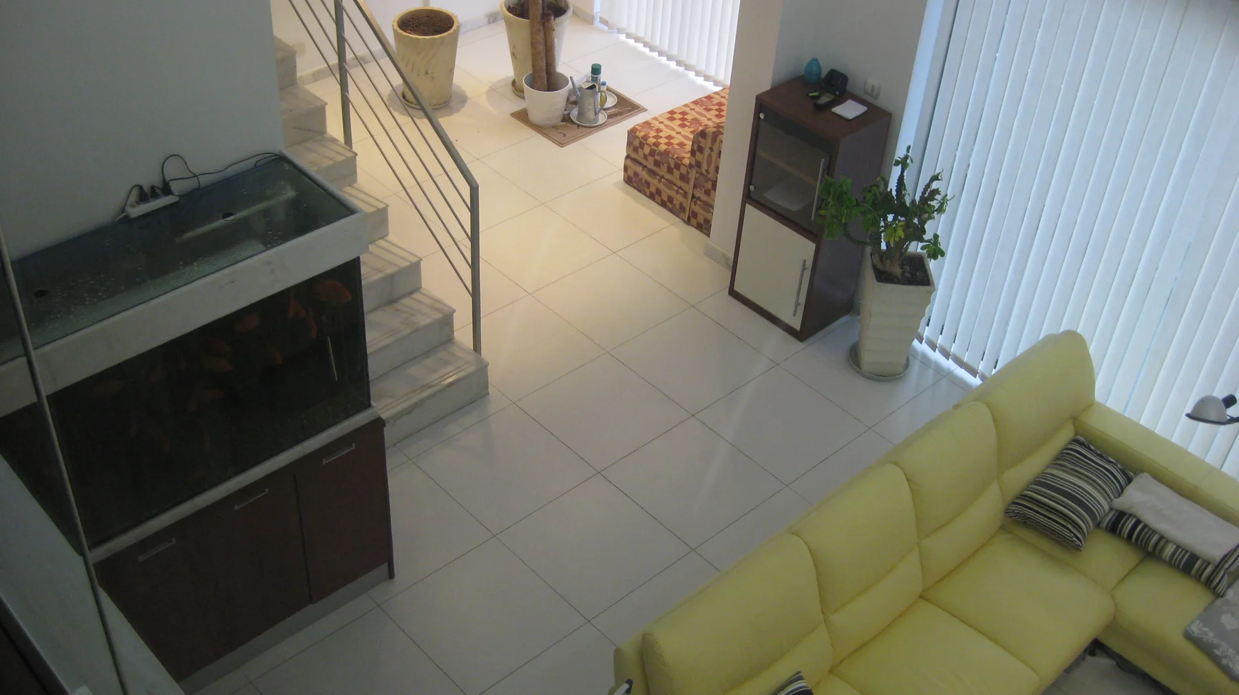 An elevated indoor view shows a pale sofa, tiled floor, dark shelving and plants beside tall windows.