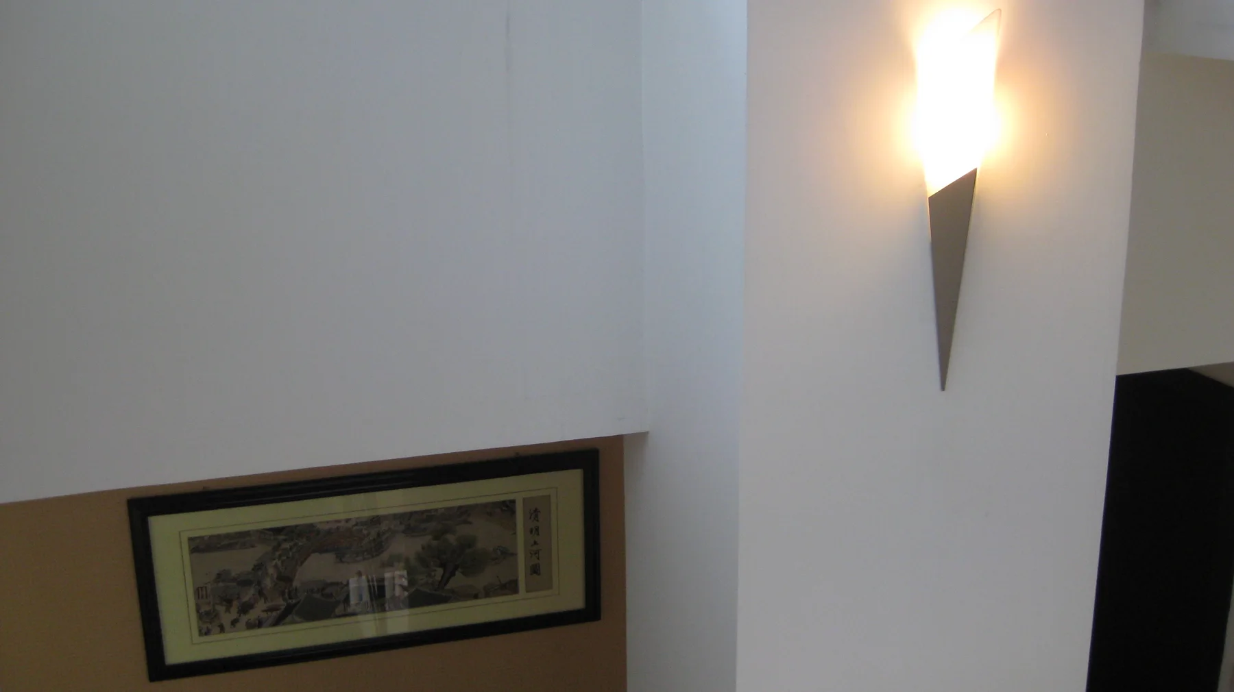 A glowing wall light hangs above and beside a framed picture on a pale interior wall.